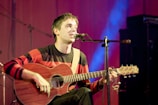 A person is seated on a stage, playing an acoustic guitar and singing into a microphone. They are wearing a red and black striped sweater, and the background is illuminated with soft blue and purple lighting. The atmosphere suggests a live music performance.
