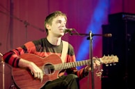A person is seated on a stage, playing an acoustic guitar and singing into a microphone. They are wearing a red and black striped sweater, and the background is illuminated with soft blue and purple lighting. The atmosphere suggests a live music performance.