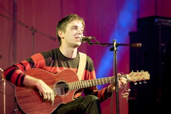 A person is seated on a stage, playing an acoustic guitar and singing into a microphone. They are wearing a red and black striped sweater, and the background is illuminated with soft blue and purple lighting. The atmosphere suggests a live music performance.