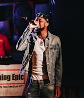 A man is performing on stage, holding a microphone close to his mouth. He wears a denim jacket over a white shirt, a cap with patterns, and a pendant necklace. There is a backdrop with a partially blurred image and text that reads 'Epic'. Another person in red is partially visible in the background.