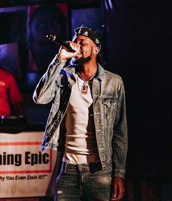 A man is performing on stage, holding a microphone close to his mouth. He wears a denim jacket over a white shirt, a cap with patterns, and a pendant necklace. There is a backdrop with a partially blurred image and text that reads 'Epic'. Another person in red is partially visible in the background.