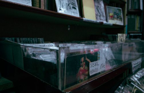 A dimly lit shelf filled with various records or CDs in transparent cases. One case has a label saying 'Special Hawaii' priced at 15€. The shelf is surrounded by other stacks of media, possibly books or albums.