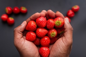 Hands are gently cupping a pile of fresh, ripe strawberries against a dark background. A few more strawberries are scattered in the background, enhancing the vividness of the red fruits.