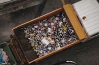 A large container is filled with various types of cans and bottles, mostly metal, which appear to be recyclable waste. It is placed on the back of a truck, positioned on a grey concrete surface.