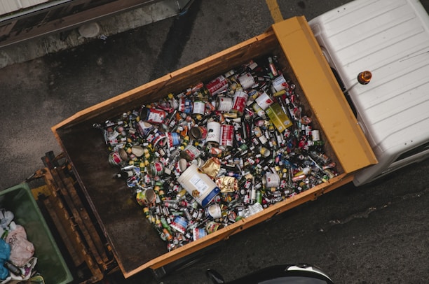 A large container is filled with various types of cans and bottles, mostly metal, which appear to be recyclable waste. It is placed on the back of a truck, positioned on a grey concrete surface.