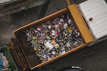 A large container is filled with various types of cans and bottles, mostly metal, which appear to be recyclable waste. It is placed on the back of a truck, positioned on a grey concrete surface.