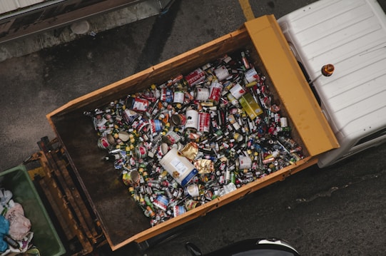A large container is filled with various types of cans and bottles, mostly metal, which appear to be recyclable waste. It is placed on the back of a truck, positioned on a grey concrete surface.