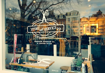 A stylish storefront window features a logo and text advertising a store specialized in repairs, alterations, laundry, and coffee to go. The reflection of a scenic urban street with historic buildings can be seen in the glass, blending the interior workspace with vintage sewing machines and tools, and the outside world.