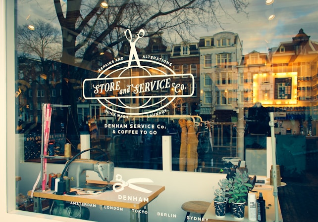 A stylish storefront window features a logo and text advertising a store specialized in repairs, alterations, laundry, and coffee to go. The reflection of a scenic urban street with historic buildings can be seen in the glass, blending the interior workspace with vintage sewing machines and tools, and the outside world.