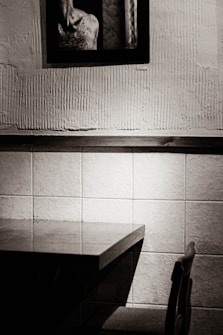 A monochromatic photograph depicts a minimalist interior scene featuring an empty table with a single chair beside it. The wall in the background is textured and divided into two sections, with the lower part tiled and the upper part featuring rough plaster. A framed picture hangs in the upper section, showing part of a person's back and shoulders. The lighting is soft, casting subtle shadows.