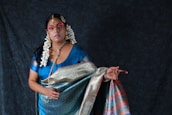 A woman dressed in traditional attire featuring a richly patterned sari in shades of blue, green, pink, and gold. She is adorned with elaborate jewelry, including large earrings and a headpiece. Her hair is styled with white flowers, and her face is painted with makeup that highlights her eyes and lips. The background is a dark, textured fabric, enhancing the vibrancy of her clothing.