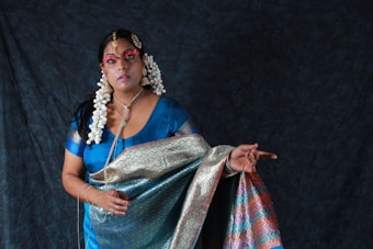 A woman dressed in traditional attire featuring a richly patterned sari in shades of blue, green, pink, and gold. She is adorned with elaborate jewelry, including large earrings and a headpiece. Her hair is styled with white flowers, and her face is painted with makeup that highlights her eyes and lips. The background is a dark, textured fabric, enhancing the vibrancy of her clothing.