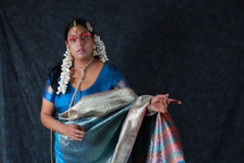 A woman dressed in traditional attire featuring a richly patterned sari in shades of blue, green, pink, and gold. She is adorned with elaborate jewelry, including large earrings and a headpiece. Her hair is styled with white flowers, and her face is painted with makeup that highlights her eyes and lips. The background is a dark, textured fabric, enhancing the vibrancy of her clothing.