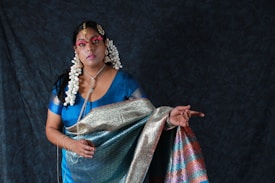 A woman dressed in traditional attire featuring a richly patterned sari in shades of blue, green, pink, and gold. She is adorned with elaborate jewelry, including large earrings and a headpiece. Her hair is styled with white flowers, and her face is painted with makeup that highlights her eyes and lips. The background is a dark, textured fabric, enhancing the vibrancy of her clothing.