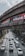 Heavy traffic with cars and motorcycles moving on a multi-lane road beneath an elevated railway or bridge structure. Advertising billboards are seen on the bridge, and tall buildings and trees line the sides of the road. The sky is partly cloudy.