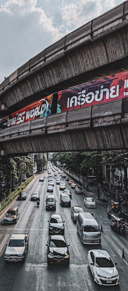 Heavy traffic with cars and motorcycles moving on a multi-lane road beneath an elevated railway or bridge structure. Advertising billboards are seen on the bridge, and tall buildings and trees line the sides of the road. The sky is partly cloudy.