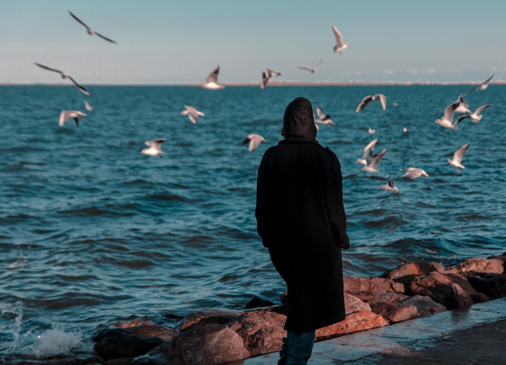 woman in black dress standing on rocky shore looking at birds flying over the sea during