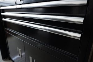 A black metal cabinet with several horizontal shiny metal handles. The cabinet appears to have multiple drawers and lower storage doors. The smooth surface reflects some light, giving it a polished appearance.