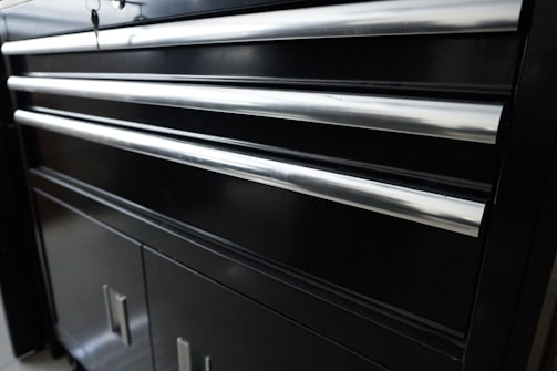 A black metal cabinet with several horizontal shiny metal handles. The cabinet appears to have multiple drawers and lower storage doors. The smooth surface reflects some light, giving it a polished appearance.