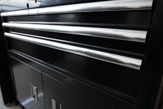 A black metal cabinet with several horizontal shiny metal handles. The cabinet appears to have multiple drawers and lower storage doors. The smooth surface reflects some light, giving it a polished appearance.