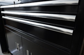A black metal cabinet with several horizontal shiny metal handles. The cabinet appears to have multiple drawers and lower storage doors. The smooth surface reflects some light, giving it a polished appearance.