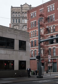 Urban street scene with buildings of various architectural styles. The foreground features a modern, concrete building while the background showcases older, brick structures with numerous windows and a visible fire escape. A person in a suit is walking on the sidewalk below, near a traffic pole and a no-turn traffic sign.