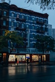 A multi-story building with a blue facade and red accents houses a hardware store on the ground floor. The signage reads 'Wankel's Hardware' and 'Paints.' The scene is set in the evening, with the interior of the store brightly illuminated, casting warm light onto the pavement. Two people with umbrellas walk along the wet street, suggesting recent rainfall. The overcast sky and dim lighting create a serene atmosphere.