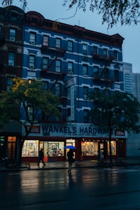A multi-story building with a blue facade and red accents houses a hardware store on the ground floor. The signage reads 'Wankel's Hardware' and 'Paints.' The scene is set in the evening, with the interior of the store brightly illuminated, casting warm light onto the pavement. Two people with umbrellas walk along the wet street, suggesting recent rainfall. The overcast sky and dim lighting create a serene atmosphere.