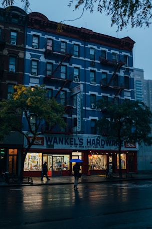 A multi-story building with a blue facade and red accents houses a hardware store on the ground floor. The signage reads 'Wankel's Hardware' and 'Paints.' The scene is set in the evening, with the interior of the store brightly illuminated, casting warm light onto the pavement. Two people with umbrellas walk along the wet street, suggesting recent rainfall. The overcast sky and dim lighting create a serene atmosphere.