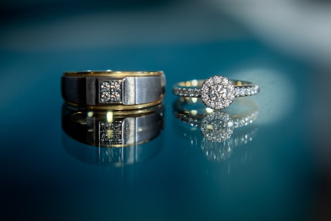 Two rings, one gold band with a rectangular design set with small diamonds and the other a diamond ring with a circular cluster on a reflective surface, showcasing their brilliance against a soft blue background.