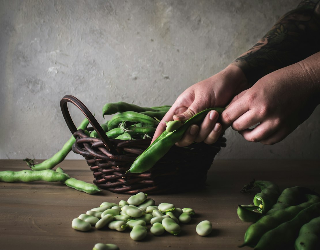 Hands shelling fresh fava beans at a wooden table