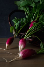 A dark brown wicker basket filled with freshly harvested radishes with green leaves spilling over onto a wooden surface. Some radishes are scattered outside the basket, showing their long roots and vibrant pink skin.
