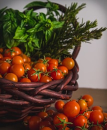 A woven basket filled with vibrant cherry tomatoes. Fresh basil leaves and rosemary sprigs are placed in the background. The scene conveys a rustic and fresh garden-to-table aesthetic.