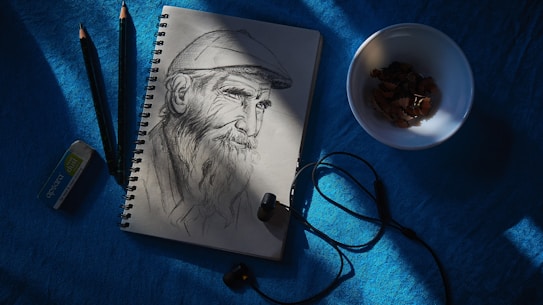 A sketch of an elderly man with a beard wearing a cap is drawn on a spiral-bound notepad. Two pencils and an eraser are placed beside the notepad, while a pair of black earbuds with a cable lie partially over the notepad. A white bowl containing pencil shavings is situated to the right.