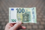 A person is holding a 100-euro banknote. The banknote features intricate architectural designs and European symbols, with shades of green and blue. The background is blurred, focusing attention on the currency.