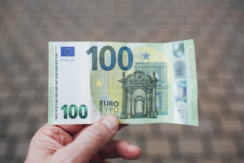 A person is holding a 100-euro banknote. The banknote features intricate architectural designs and European symbols, with shades of green and blue. The background is blurred, focusing attention on the currency.