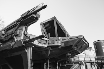 A monochrome image featuring a large, futuristic, and industrial-looking machine with detailed mechanical structures. The machine has prominent cylindrical elements and angular designs, suggesting advanced technology or a military apparatus.