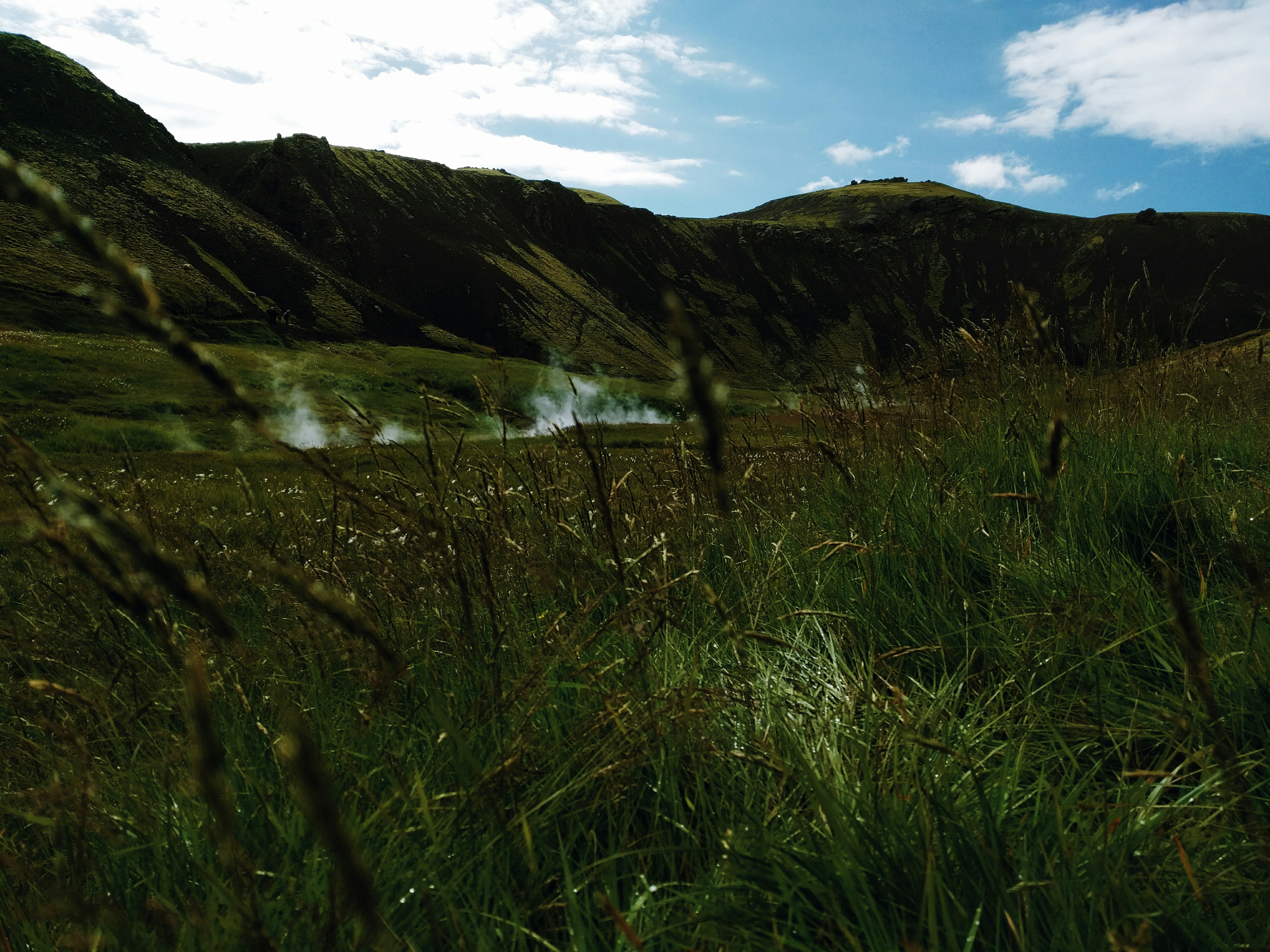 green grass field near lake under white clouds and blue sky during daytime, Reykjadalur "Steam Valley", Iceland. Olive-green hills, wildflowers, hot springs, steam, and sulfur.