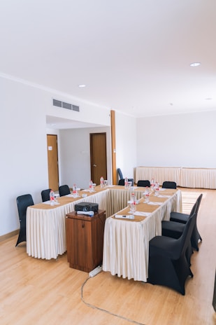 A conference room setup with a U-shaped table arrangement draped in white tablecloths. Black chairs surround the tables, each place setting includes a bottle of water and a glass. A small wooden podium or stand holds a projector and a remote, with a visible cable extending from it. The room has plain white walls and light wooden flooring, with several doors in the background.