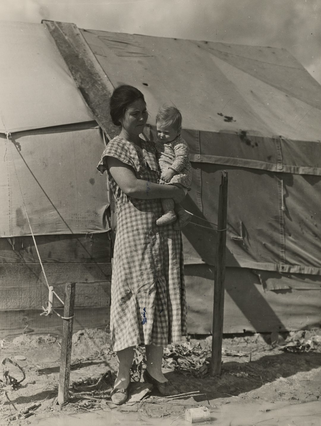 grayscale photo of woman in checked dress carrying her child while standing beside white tent