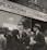 A group of men stand outside a building with a sign above them that reads 'World War Veterans applications for the bonus filled out here'. The men are dressed in suits and coats, with some wearing hats, indicative of a mid-20th century style. They appear to be engaged in conversation, with one man gesturing as he speaks. The background shows a brick building with large windows.