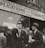 A group of men stand outside a building with a sign above them that reads 'World War Veterans applications for the bonus filled out here'. The men are dressed in suits and coats, with some wearing hats, indicative of a mid-20th century style. They appear to be engaged in conversation, with one man gesturing as he speaks. The background shows a brick building with large windows.