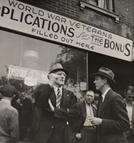 A group of men stand outside a building with a sign above them that reads 'World War Veterans applications for the bonus filled out here'. The men are dressed in suits and coats, with some wearing hats, indicative of a mid-20th century style. They appear to be engaged in conversation, with one man gesturing as he speaks. The background shows a brick building with large windows.