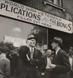 A group of men stand outside a building with a sign above them that reads 'World War Veterans applications for the bonus filled out here'. The men are dressed in suits and coats, with some wearing hats, indicative of a mid-20th century style. They appear to be engaged in conversation, with one man gesturing as he speaks. The background shows a brick building with large windows.