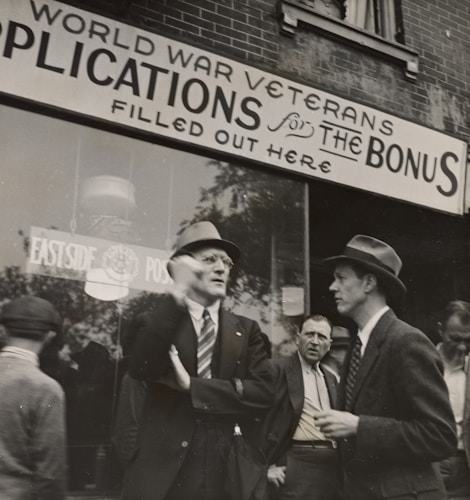 A group of men stand outside a building with a sign above them that reads 'World War Veterans applications for the bonus filled out here'. The men are dressed in suits and coats, with some wearing hats, indicative of a mid-20th century style. They appear to be engaged in conversation, with one man gesturing as he speaks. The background shows a brick building with large windows.