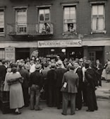 A large crowd of people stands gathered outside a brick building with signs for veteran bonus applications and music services. Many individuals wear hats and coats typical of the early 20th century. Some people can be seen on the balconies and looking out from windows, observing the crowd below.