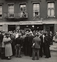 A large crowd of people stands gathered outside a brick building with signs for veteran bonus applications and music services. Many individuals wear hats and coats typical of the early 20th century. Some people can be seen on the balconies and looking out from windows, observing the crowd below.