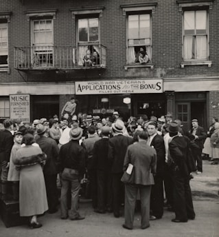 A large crowd of people stands gathered outside a brick building with signs for veteran bonus applications and music services. Many individuals wear hats and coats typical of the early 20th century. Some people can be seen on the balconies and looking out from windows, observing the crowd below.