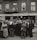A large crowd of people stands gathered outside a brick building with signs for veteran bonus applications and music services. Many individuals wear hats and coats typical of the early 20th century. Some people can be seen on the balconies and looking out from windows, observing the crowd below.