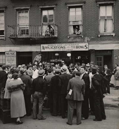 A large crowd of people stands gathered outside a brick building with signs for veteran bonus applications and music services. Many individuals wear hats and coats typical of the early 20th century. Some people can be seen on the balconies and looking out from windows, observing the crowd below.